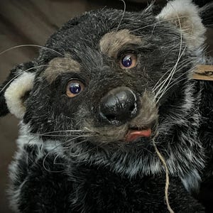 May include: A handmade plush toy in the shape of a raccoon. The toy has black and gray fur, with tan markings around the eyes and ears. It has a black nose, whiskers, and a small red tongue. The toy is detailed and appears to be made of high-quality materials.