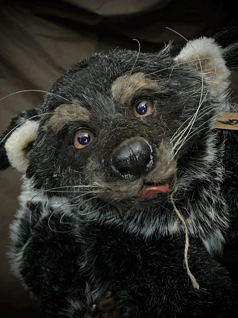 May include: A handmade plush toy in the shape of a raccoon. The toy has black and gray fur, with tan markings around the eyes and ears. It has a black nose, whiskers, and a small red tongue. The toy is detailed and appears to be made of high-quality materials.