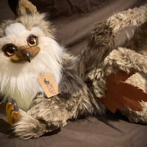 Puede incluir: Un juguete de peluche con pelaje marr&oacute;n y blanco, ojos grandes y un pico. El juguete tiene un ala de cuero marr&oacute;n y una etiqueta que dice "Krafcola Creations".