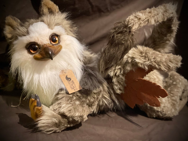 Puede incluir: Un juguete de peluche con pelaje marr&oacute;n y blanco, ojos grandes y un pico. El juguete tiene un ala de cuero marr&oacute;n y una etiqueta que dice "Krafcola Creations".