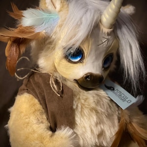 Puede incluir: Un unicornio de peluche blanco y marrón con un cuerno largo y blanco, ojos azules y plumas marrones. El unicornio lleva un chaleco marrón y tiene una etiqueta que dice "HREAFLO ARTISANS".
