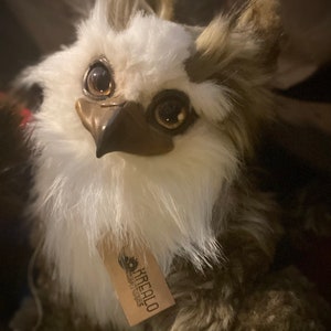 Puede incluir: Un peque&ntilde;o peluche de b&uacute;ho marr&oacute;n y blanco, suave y esponjoso, con grandes ojos dorados. El juguete tiene un pico marr&oacute;n y una etiqueta que dice "Kirealo Creations".