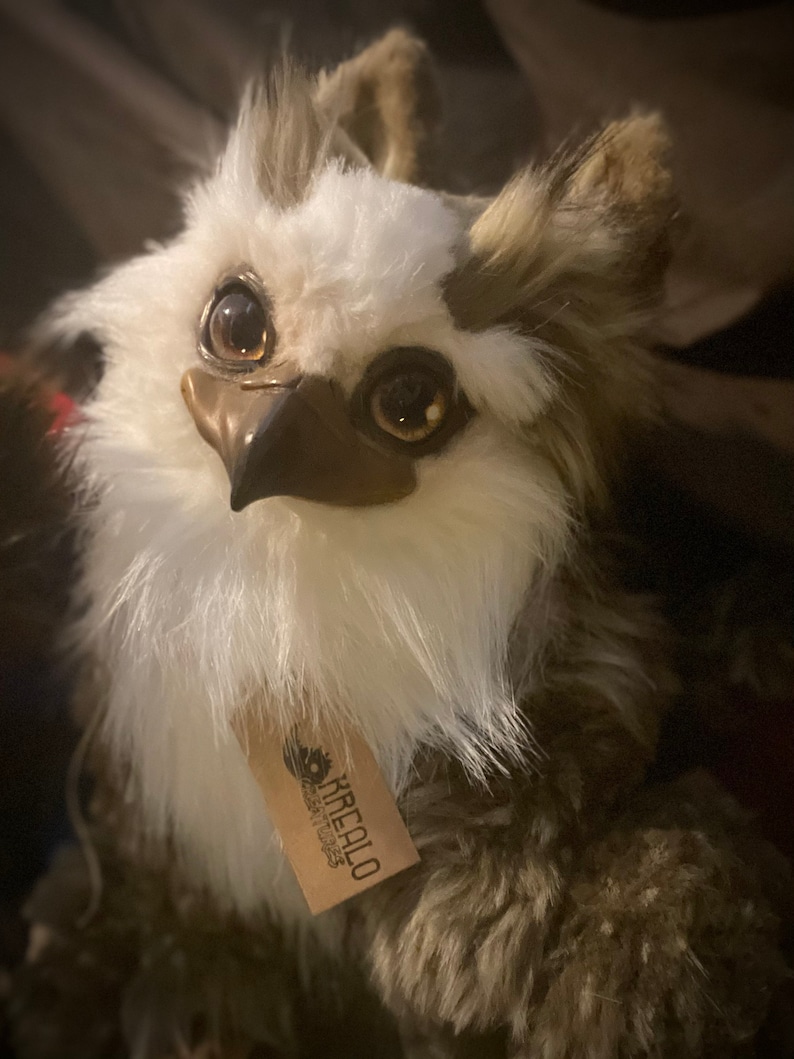 Puede incluir: Un peque&ntilde;o peluche de b&uacute;ho marr&oacute;n y blanco, suave y esponjoso, con grandes ojos dorados. El juguete tiene un pico marr&oacute;n y una etiqueta que dice "Kirealo Creations".