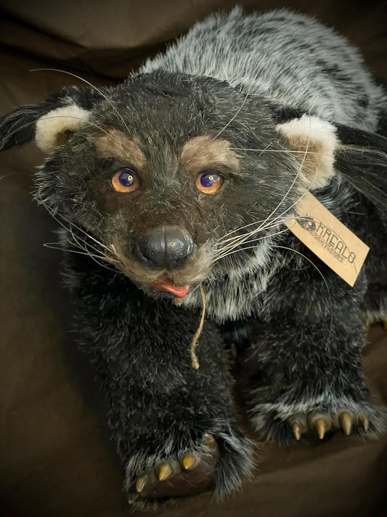 May include: A handmade plush animal with a black and gray fur coat, amber eyes, and a small pink tongue. The plush has a tag attached with the name "KREALA". The animal's paws have yellow claws.