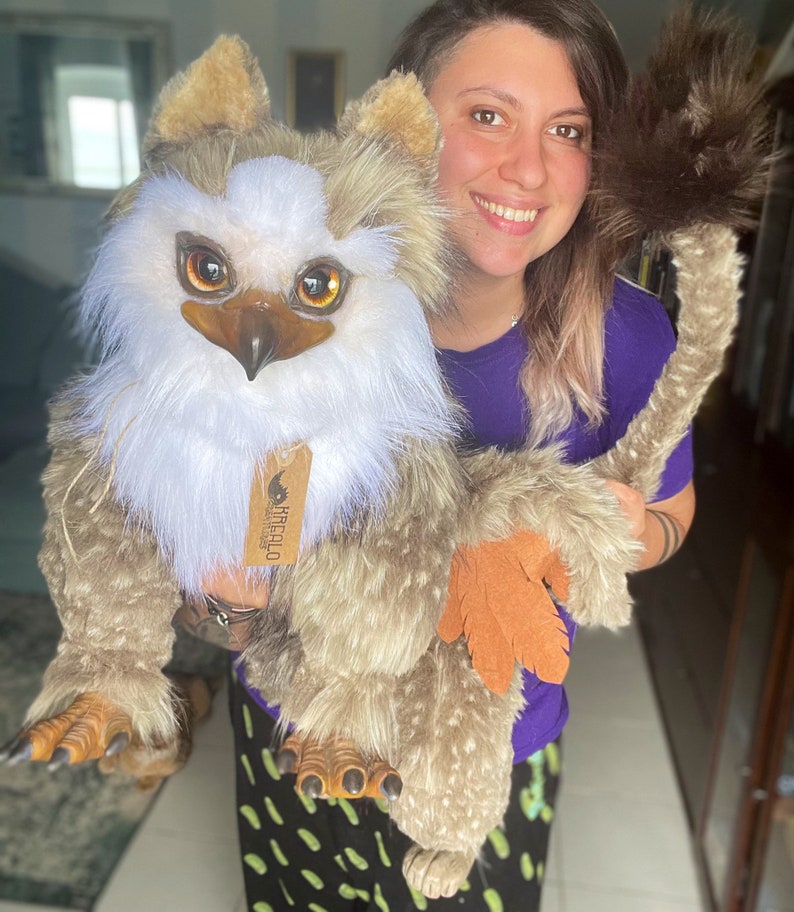 Puede incluir: Una persona sosteniendo un gran juguete de peluche marr&oacute;n y blanco con una cola larga. El juguete tiene una cara de p&aacute;jaro con ojos grandes de color naranja y un pico. El juguete tiene una etiqueta que dice "Harecalo Creations".