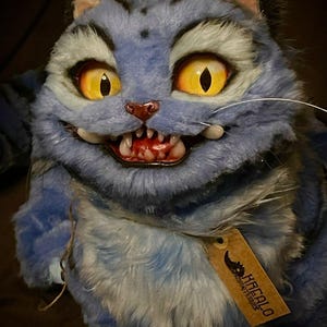 Puede incluir: Un gato de peluche azul con una amplia sonrisa, ojos amarillos y dientes visibles. El gato tiene una apariencia de pelaje detallada y texturizada. Una pequeña etiqueta con el texto "KREALO ADVENTURES" está adjunta.