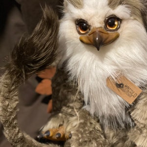 Puede incluir: Un juguete de peluche con pelaje marr&oacute;n y blanco, un pico de p&aacute;jaro y grandes ojos dorados. El juguete tiene una cola larga y tupida y est&aacute; sentado sobre una superficie marr&oacute;n. El juguete tiene una etiqueta que dice "KREALO DESIGN STUDIO".