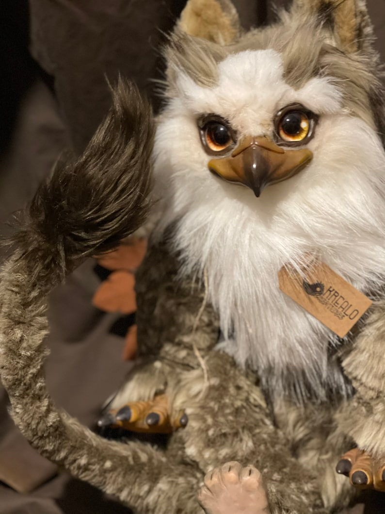 Puede incluir: Un juguete de peluche con pelaje marr&oacute;n y blanco, un pico de p&aacute;jaro y grandes ojos dorados. El juguete tiene una cola larga y tupida y est&aacute; sentado sobre una superficie marr&oacute;n. El juguete tiene una etiqueta que dice "KREALO DESIGN STUDIO".