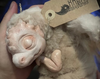 Puede incluir: Un pequeño dragón de peluche blanco con los ojos cerrados y una cara esculpida texturizada. El dragón está acostado de lado con la cabeza apoyada en su brazo. El dragón está etiquetado con una etiqueta de papel marrón que dice "KREALO CREATURES".