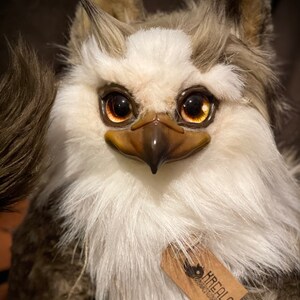 Puede incluir: Un juguete de peluche blanco y marr&oacute;n con un pico y ojos grandes y dorados. El juguete tiene una etiqueta que dice "KREALO Creations".