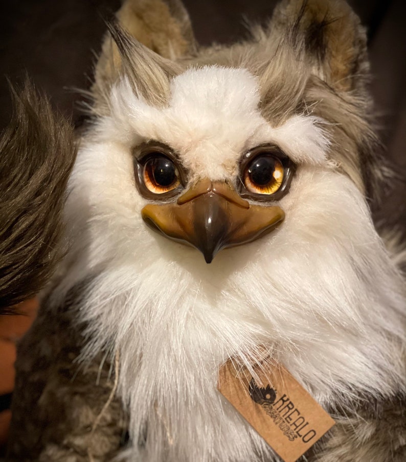 Puede incluir: Un juguete de peluche blanco y marr&oacute;n con un pico y ojos grandes y dorados. El juguete tiene una etiqueta que dice "KREALO Creations".