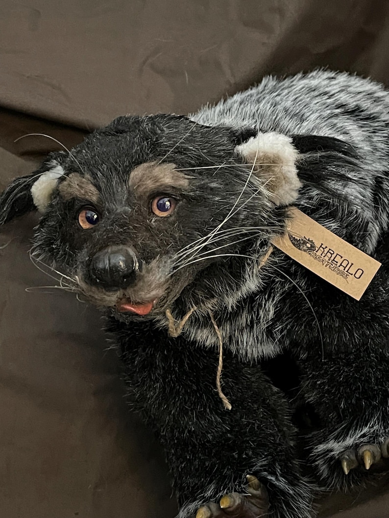 May include: A plush, lifelike badger toy with black, gray, and tan fur. The toy has a detailed face with pink eyes, a black nose, and long white whiskers. A small tag is attached to the toy.