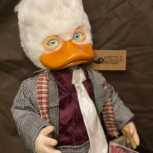 Könnte beinhalten: Eine weiße Plüschpuppe mit einem Entenkopf, die einen grauen karierten Anzug mit einem burgunderfarbenen Hemd und einer weißen Krawatte trägt. Die Puppe hält ein Magazin mit dem Text "Fighting Egg" auf dem Cover.