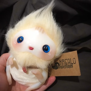 Könnte beinhalten: Weißes, flauschiges Wesen mit blauen Augen und einem roten Mund. Das Wesen hält ein weißes Tuch in seinen Händen. Das Wesen sitzt auf einer Hand. Ein braunes Etikett mit dem Text "KREALO CREATURES" ist im Hintergrund sichtbar.
