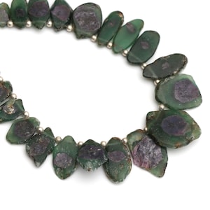 May include: A necklace featuring irregularly shaped, translucent green beads with dark purple centers. The beads are strung together with small silver-colored beads. The necklace is displayed against a white background.