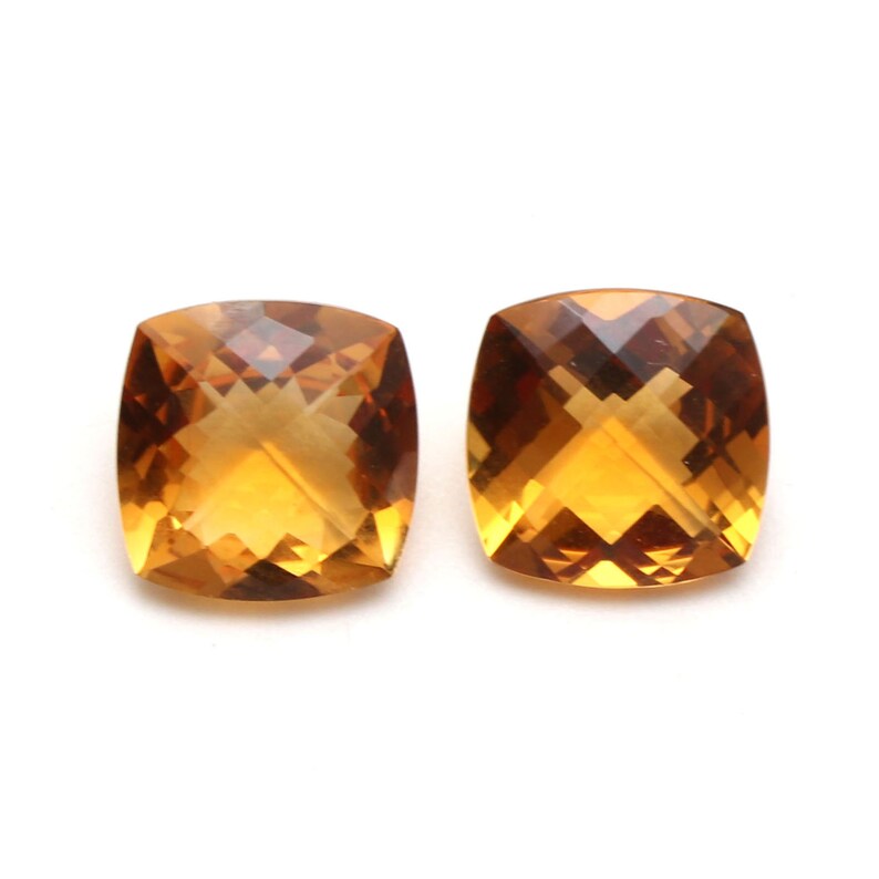 May include: Two cushion-cut citrine gemstones, each with a warm, golden-amber hue. The stones are faceted and have a high degree of brilliance.