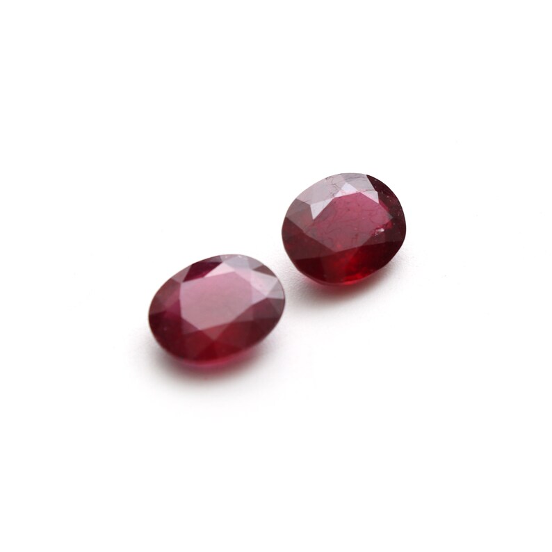 May include: Two oval-shaped, red gemstones with a faceted cut. The gemstones are set against a white background.