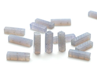 Puede incluir: Un conjunto de cuentas rectangulares de piedra preciosa azul pálido con un patrón geométrico y agujeros perforados. Las cuentas están dispersas sobre una superficie blanca, mostrando su cualidad translúcida y su potencial para la fabricación de joyas.