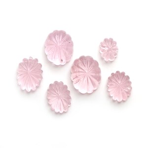 May include: A collection of six light pink gemstone flower-shaped carvings. The carvings have a detailed petal design radiating from the center. The gemstones are arranged on a white background, showcasing their delicate color and intricate details.