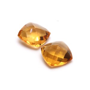 May include: Two cushion-cut topaz gemstones, one slightly larger than the other, are displayed on a white background. The gemstones are a light to medium shade of yellow.