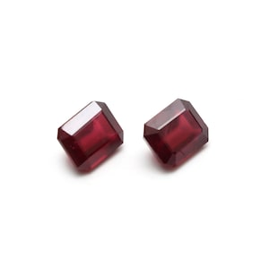 May include: Two red emerald cut gemstones on a white background.