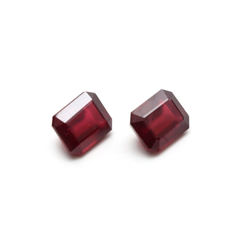 May include: Two red emerald cut gemstones on a white background.