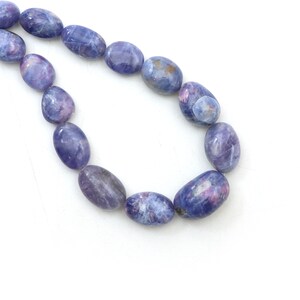 Puede incluir: Una hebra de cuentas de piedras preciosas ovaladas de color azul y morado. Las cuentas tienen un aspecto moteado y nublado.