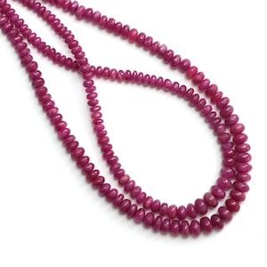 May include: Strands of polished, deep pink ruby beads. The beads are small, uniform in size, and strung together to form necklaces. The background is plain white, highlighting the vibrant color and texture of the gemstones. The necklaces are displayed in a layered arrangement.