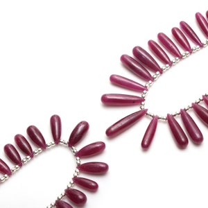 Może przedstawiać: Szereg koralików z rubinowych kamieni szlachetnych nawleczonych na srebrny łańcuszek. Koraliki mają kształt łzy i są polerowane.