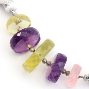 May include: A close-up of a necklace with alternating light green, purple, and pink gemstone beads. The beads are faceted and have a smooth, polished finish. The necklace is strung on a silver chain.