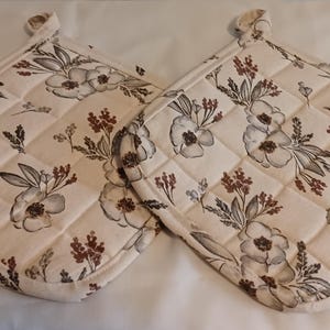 May include: Two quilted pot holders with a floral pattern. The pot holders are cream-colored with a repeating design of gray and brown flowers and leaves. Each pot holder has a loop for hanging.