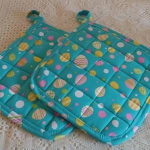 May include: Two turquoise quilted pot holders with an Easter egg pattern. The pattern includes white, pink, and yellow eggs. Each pot holder has a loop for hanging.