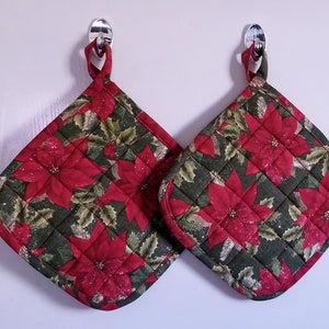 May include: Two red and green quilted pot holders with a poinsettia pattern. The pot holders are hanging on hooks.