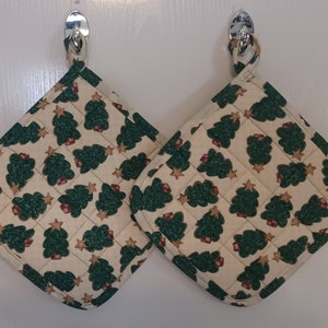 May include: Two quilted pot holders with a cream background and a repeating pattern of green Christmas trees and gold stars. The pot holders are square with a loop for hanging.