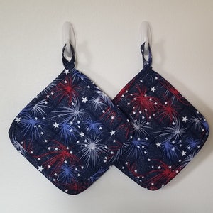 May include: Two blue quilted pot holders with a red, white, and blue firework pattern. The pot holders are hanging on hooks.