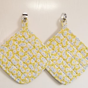 May include: Two yellow quilted pot holders with a white daisy pattern. The pot holders have a loop handle and are hanging on hooks.