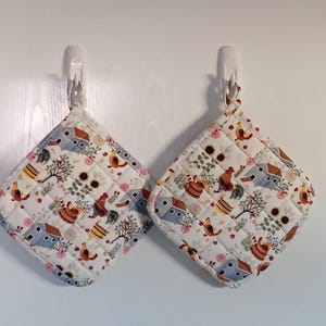 May include: Two quilted pot holders with a farm-themed pattern. The fabric features images of chickens, houses, trees, and sunflowers in shades of brown, blue, and pink on a white background. The pot holders are diamond-shaped and have a white binding.