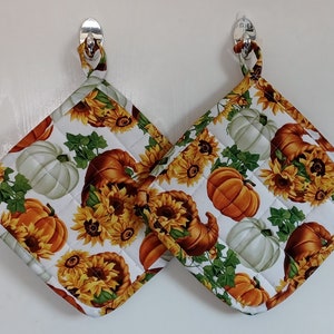 May include: Two white and green pot holders with a fall theme. The pot holders feature pumpkins, sunflowers, and fall leaves.