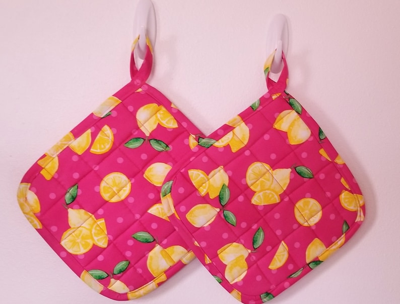 May include: Two pink quilted hot pads with a lemon print and white polka dots. The hot pads have a loop for hanging.