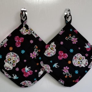 May include: Two black quilted pot holders with a sugar skull design. The skulls are decorated with pink, blue, and white details. The pot holders have a loop for hanging.