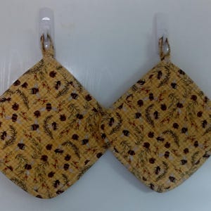 May include: Two yellow quilted pot holders with a repeating pattern of bees, pinecones, and foliage. The pot holders are square with rounded corners and a loop for hanging. The background is a white wall.