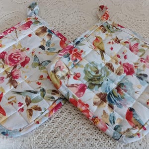 May include: Two white quilted pot holders with a floral and bird print. The print features pink, blue, and yellow flowers, and small birds.