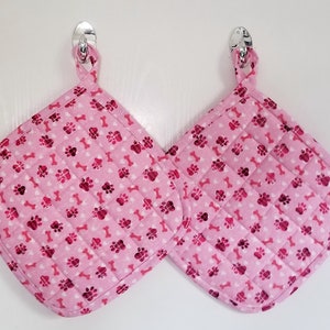 May include: Two pink quilted pot holders with a white background and a pattern of pink paw prints and white bones.
