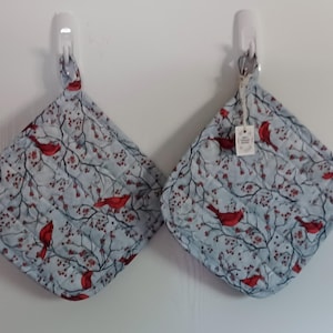 May include: Two square quilted potholders featuring a light blue background with a pattern of red cardinal birds perched on dark brown branches with small red berries. Each potholder has a fabric loop for hanging and one has a small white tag.