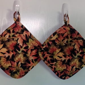 May include: Two quilted pot holders with a loop for hanging. The pot holders feature a fall leaf pattern in shades of red, orange, yellow, and green on a black background. The pot holders are square-shaped with a quilted design.