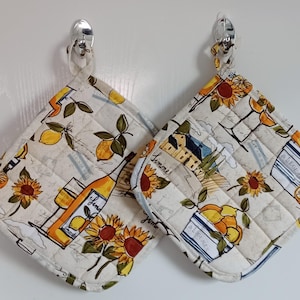 May include: Two white quilted pot holders with a lemon and sunflower design. One pot holder features a bottle of wine and a glass of wine. The other pot holder features a basket of lemons and a small house with the word "Limone" written on it.