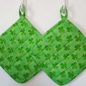 May include: Two green pot holders with a shamrock and swirl pattern. The pot holders have gold glitter accents.