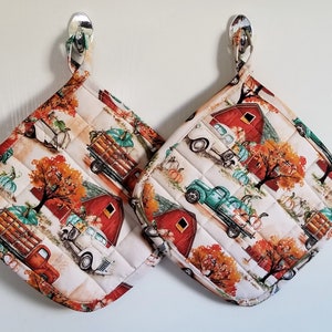 May include: Two quilted pot holders with a fall theme. The pot holders feature a white background with a red barn, fall trees, pumpkins, and vintage trucks carrying pumpkins. The trucks are teal, red, and white.