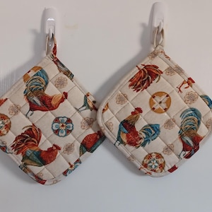 May include: Two beige quilted pot holders with a rooster and floral pattern. The pot holders are hanging on hooks.