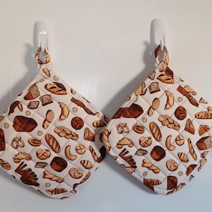 May include: Two white quilted pot holders with a brown and white pattern of various breads and pastries. The pot holders are hanging on white hooks.